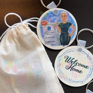 May include: A set of two round keychains with colorful watercolor backgrounds. One keychain features a cartoon illustration of a woman holding a "SOLD" sign with the text "SHELLEY AKILI REALTOR (757) 651-0585 #soldbyshelley". The other keychain features the text "Welcome Home" in blue cursive lettering.