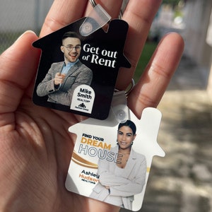 May include: Two house-shaped keychains with photos of real estate agents. The top keychain is black with white text that reads "Get out of Rent" and "Mike Smith REALTOR". The bottom keychain is white with black text that reads "Find Your Dream House" and "Ashley Hudson REALTOR".