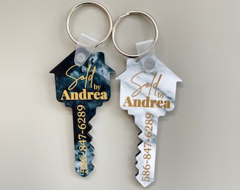 May include: Two house-shaped keychains with a key design. Both keychains are white and black with a marble pattern. The text "Sold by Andrea" and a phone number are printed in gold on each keychain.