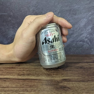 May include: A silver Asahi Super Dry beer can held by a hand. The can features Japanese text and the words "DRY" and "Asahi" in English. The background is a dark gray wall and a wooden surface.