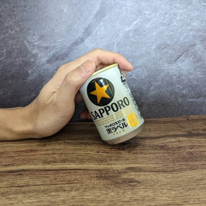 May include: A small Sapporo beer can, primarily white with black and gold accents, held by a hand. The can features the Sapporo logo with a gold star and Japanese text. The can is on a wooden surface.