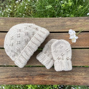 May include: A beige knitted hat and matching mittens with a textured pattern. The hat and mittens are lying on a wooden surface.