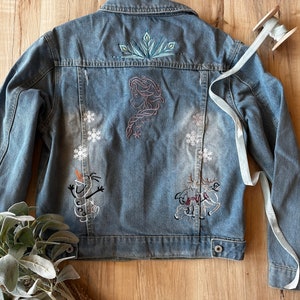 May include: A blue denim jacket with embroidered images of characters from the movie Frozen, including Elsa, Olaf, and Sven. The jacket has a distressed look with faded areas.