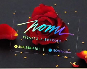 Könnte beinhalten: Eine transparente Plastik-Visitenkarte mit holografischen Goldfolien-Akzenten. Die Karte zeigt den Text "nomi PILATES + BEYOND" und Kontaktinformationen, darunter eine Telefonnummer und einen Instagram-Handle.