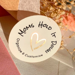 Puede incluir: Pegatina redonda blanca con letras en lámina dorada que dicen "MOMS HOLD IT DOWN" con un corazón de lámina dorada en el centro. La pegatina está sobre un fondo rosa y blanco.