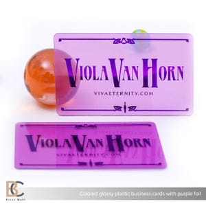 May include: Two glossy, rectangular business cards with purple foil. The cards feature the name "Viola Van Horn" and the website "VIVAETERNITY.COM" in a dark purple font. An orange glass sphere is next to the cards.