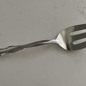 Pode incluir: Um garfo de servir prateado com três dentes e um design ranhurado. O cabo tem um padrão decorativo e ornamentado. O garfo é feito de um metal brilhante e refletor, provavelmente aço inoxidável ou prata. O utensílio foi projetado para servir alimentos.