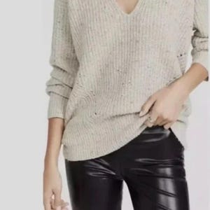 May include: A beige V-neck sweater with a ribbed knit texture and subtle multicolored flecks throughout the fabric, paired with black faux leather pants that have a smooth, shiny finish and a fitted silhouette.
