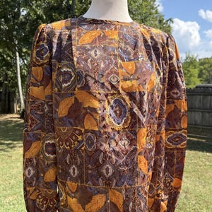 May include: A long-sleeved blouse featuring a patchwork-style print with autumn-inspired colours including mustard yellow, deep brown, and muted purple. The pattern consists of repeating paisley motifs and detailed leaf illustrations on a grid layout.