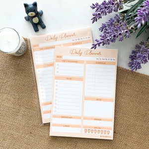 Op de afbeelding: Twee perzikkleurige dagelijkse plannerblokken met witte lijnen en cirkels om op te schrijven. Het bovenste blok heeft de tekst "Daily Planner" en het onderste blok heeft de tekst "Daily Planner". Beide blokken hebben secties voor "Date", "Top 3 Priorities", "To Do", "Schedule", "Note", "Breakfast", "Lunch" en "Dinner". Het onderste blok heeft ook een sectie voor "Water" met acht glazen.