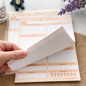 May include: A stack of peach-colored daily planner notepads with a white interior. The notepad has sections for "Date", "Top 3 Priorities", "To Do", "Schedule", "Breakfast", "Lunch", "Dinner", and "Water". The notepad also includes a line for "Mon Tue Wed Thu Fri Sat Sun".