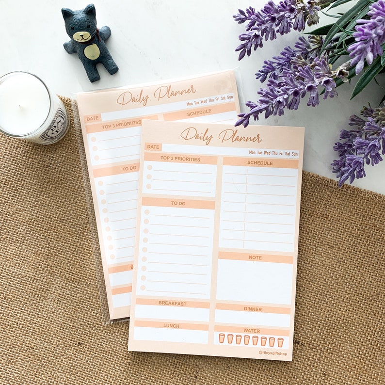May include: Two peach-colored daily planner pads with white lines and circles for writing. The top pad has the text "Daily Planner" and the bottom pad has the text "Daily Planner". Both pads have sections for "Date", "Top 3 Priorities", "To Do", "Schedule", "Note", "Breakfast", "Lunch", and "Dinner". The bottom pad also has a section for "Water" with eight glasses.