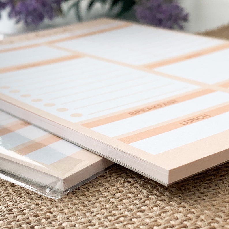 May include: A close-up of a stack of two pink and white to-do list pads. The top pad has the words "BREAKFAST" and "LUNCH" printed on it.