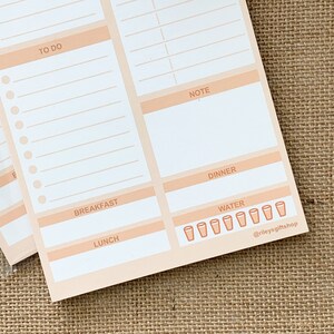 May include: A peach-colored daily planner with sections for to-do, note, breakfast, lunch, dinner, and water intake. The water intake section has eight glasses with a peach outline.