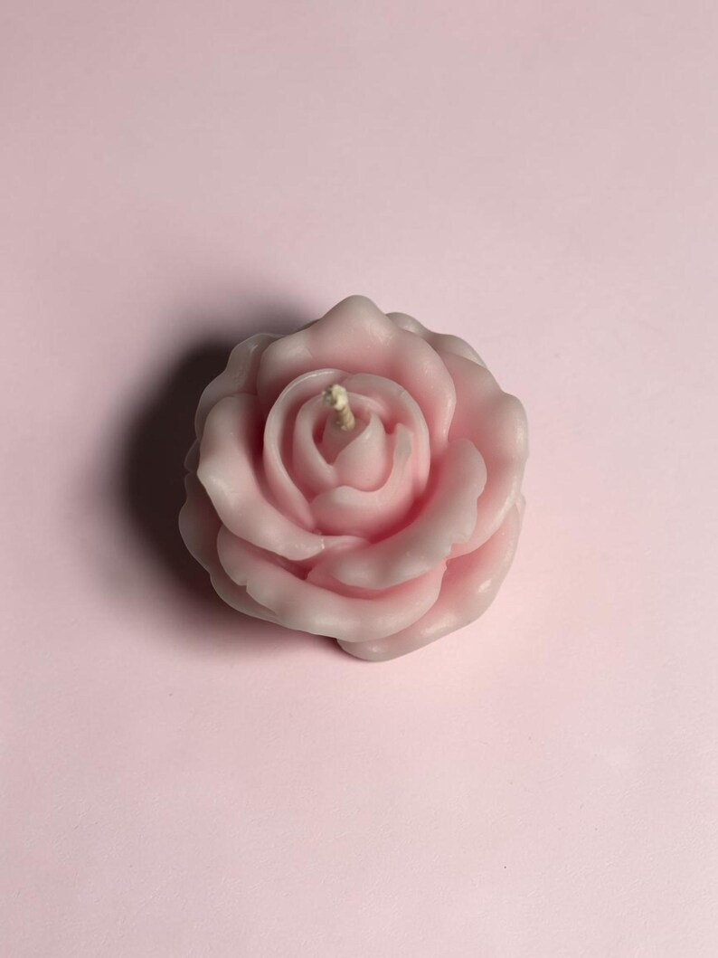 May include: A light pink candle shaped like a blooming rose flower. The wax petals are layered in a circular pattern, radiating from the center where a single, short, off-white cotton wick is positioned upright in the middle of the flower.