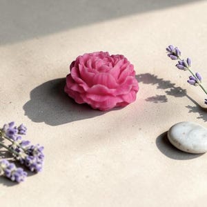 Pu&ograve; includere: Un sapone rosa acceso a forma di fiore con dettagli intricati di petali sovrapposti. Il sapone &egrave; posizionato accanto a due rametti di lavanda viola e a un singolo sasso di fiume liscio grigio chiaro, proiettando ombre nette al sole.