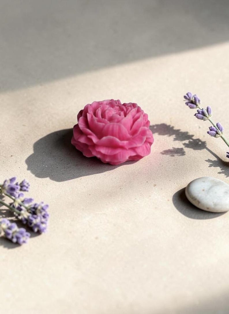Pu&ograve; includere: Un sapone rosa acceso a forma di fiore con dettagli intricati di petali sovrapposti. Il sapone &egrave; posizionato accanto a due rametti di lavanda viola e a un singolo sasso di fiume liscio grigio chiaro, proiettando ombre nette al sole.