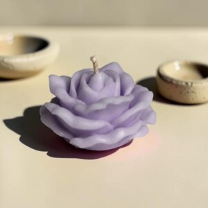 May include: A light purple candle shaped like a blooming rose with layered petals and a single central wick. The wax flower has a smooth, matte finish and is positioned between two small, textured, off-white ceramic bowls on a flat surface.