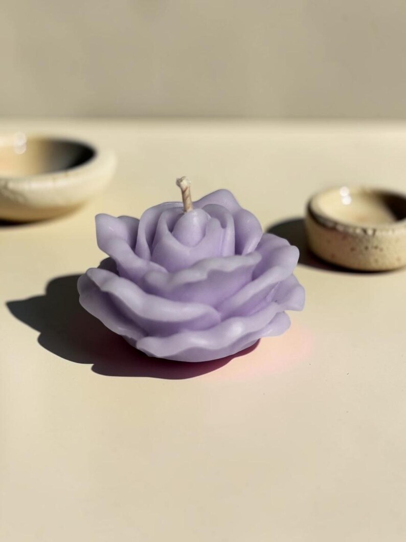 May include: A light purple candle shaped like a blooming rose with layered petals and a single central wick. The wax flower has a smooth, matte finish and is positioned between two small, textured, off-white ceramic bowls on a flat surface.