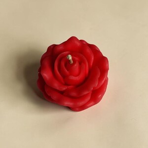 May include: A bright red wax candle shaped like a blooming rose. The candle features layered, sculpted petals that spiral toward the center, where a single, short, light-colored wick is positioned upright in the middle of the flower head.