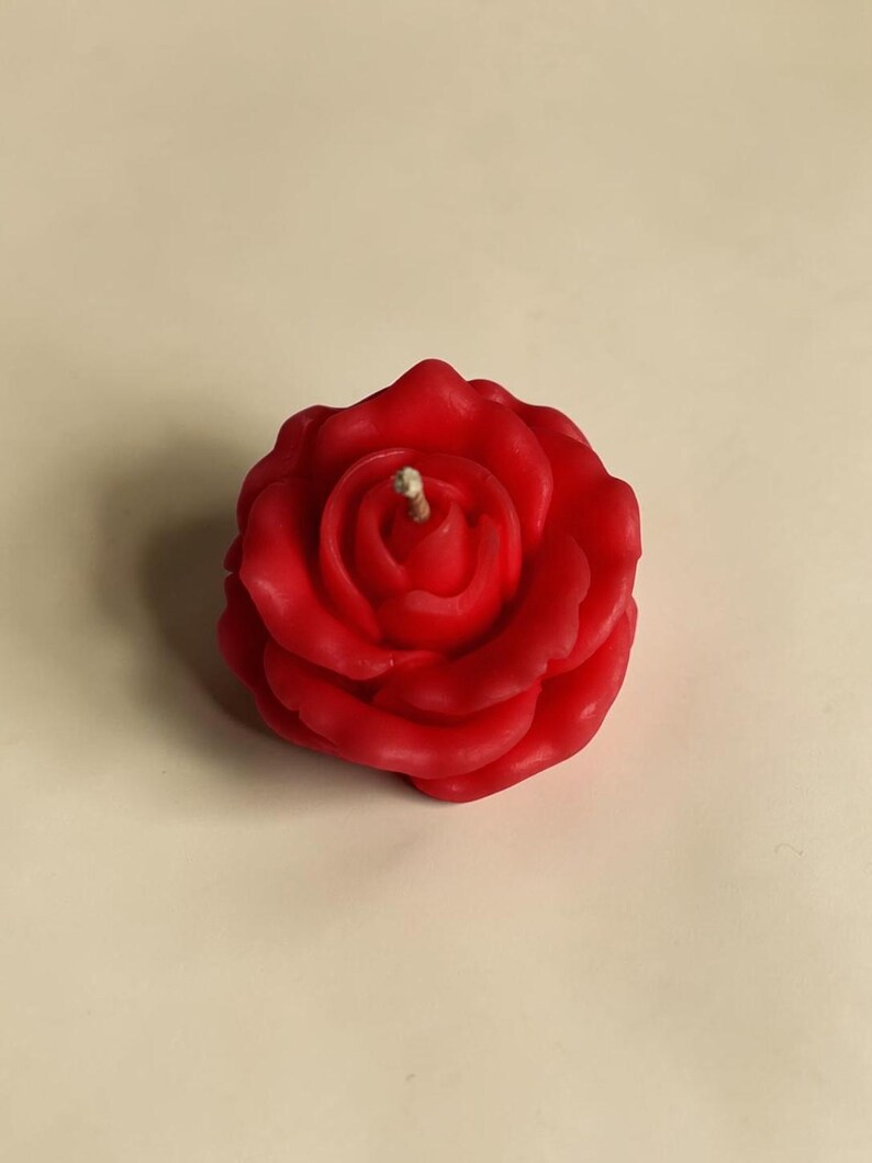 May include: A bright red wax candle shaped like a blooming rose. The candle features layered, sculpted petals that spiral toward the center, where a single, short, light-colored wick is positioned upright in the middle of the flower head.
