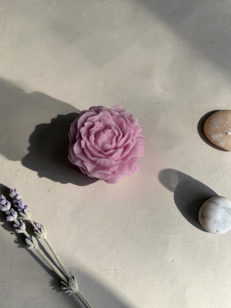 Pu&ograve; includere: Un oggetto in cera color viola chiaro modellato a forma di fiore di peonia in piena fioritura. Il fiore presenta petali complessi e stratificati. Ad accompagnare la forma floreale ci sono due piccole pietre lisce e arrotondate e un rametto di lavanda.
