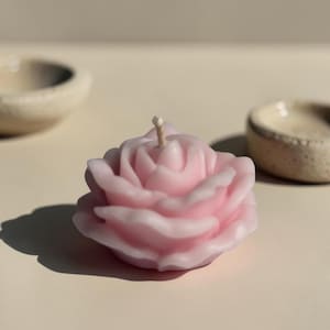 May include: A light pink rose-shaped candle with a central wick. The candle features layered, sculpted petals that mimic the appearance of a blooming flower. The wax has a smooth, matte finish and is positioned upright on a flat surface.