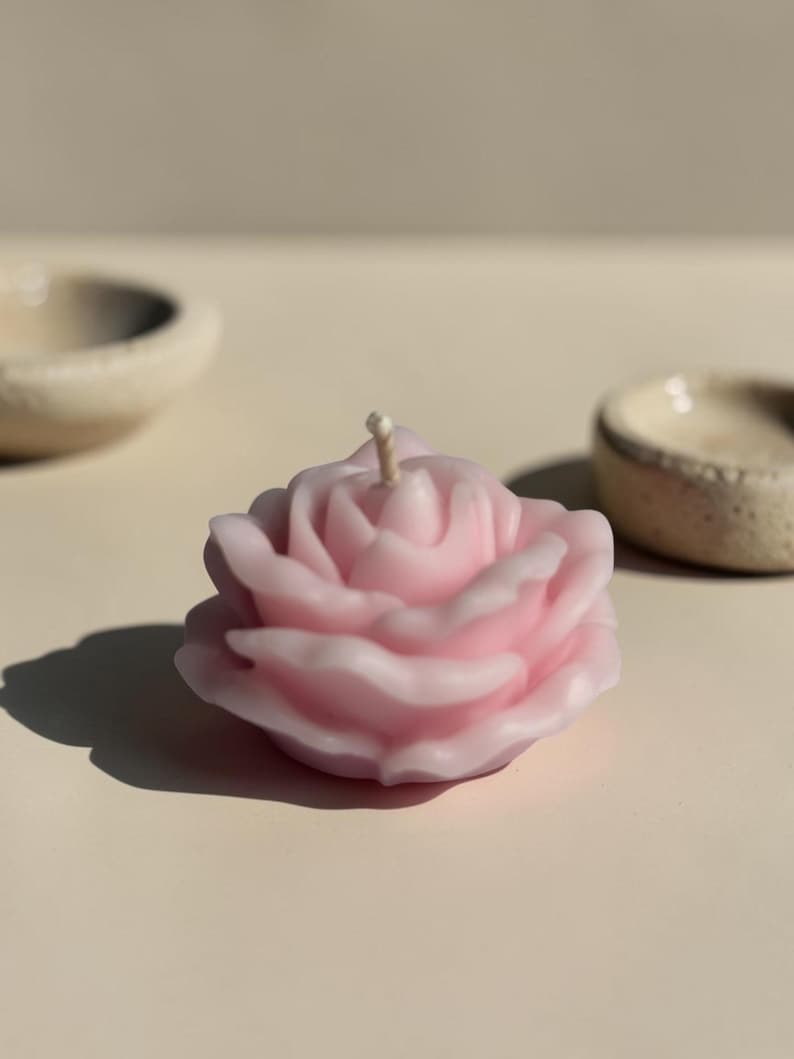 May include: A light pink rose-shaped candle with a central wick. The candle features layered, sculpted petals that mimic the appearance of a blooming flower. The wax has a smooth, matte finish and is positioned upright on a flat surface.