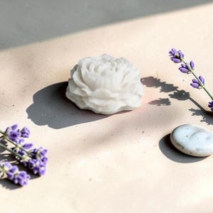 Pu&ograve; includere: Una saponetta bianca a forma di fiore con dettagli intricati di petali sovrapposti. Il sapone &egrave; posizionato accanto a due rametti di lavanda viola e a un singolo ciottolo liscio di colore grigio chiaro, creando una disposizione semplice e pulita.