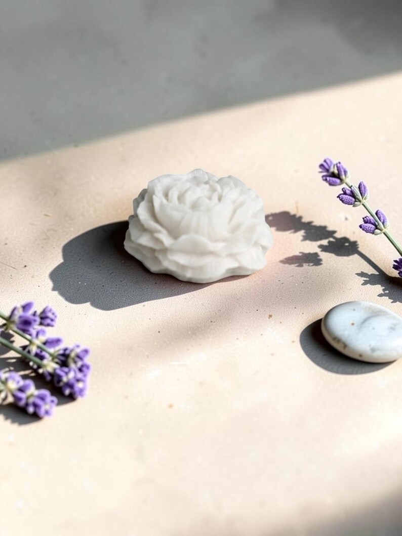 Pu&ograve; includere: Una saponetta bianca a forma di fiore con dettagli intricati di petali sovrapposti. Il sapone &egrave; posizionato accanto a due rametti di lavanda viola e a un singolo ciottolo liscio di colore grigio chiaro, creando una disposizione semplice e pulita.