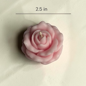 May include: A light pink rose-shaped candle with a central wick. The candle features layered, sculpted petals forming a floral bloom. A horizontal line and text indicate a measurement of 2.5 inches, showing the width of the candle from edge to edge.
