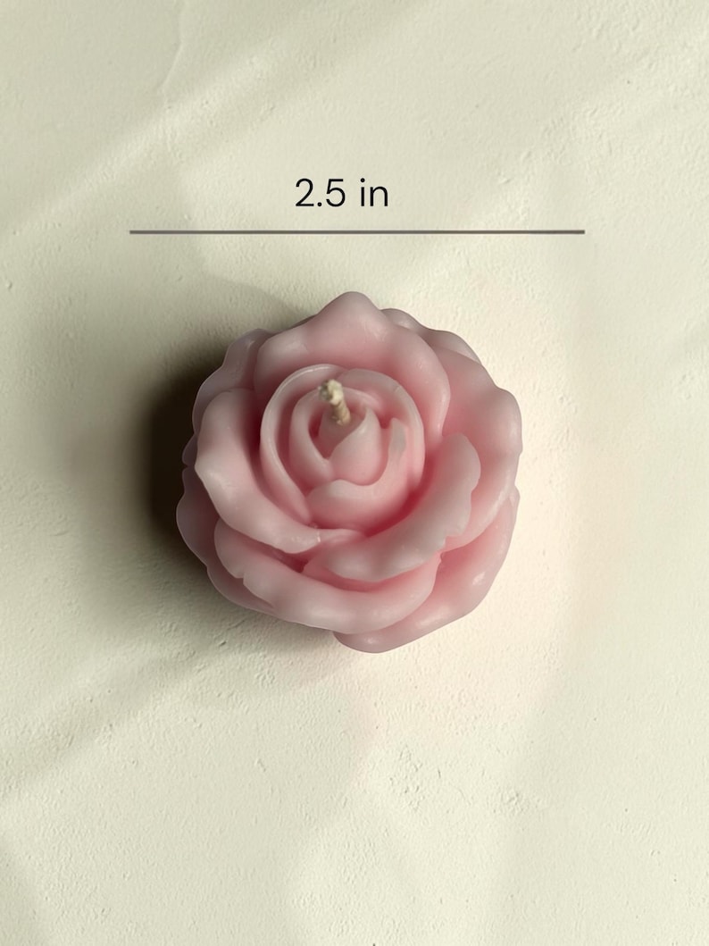 May include: A light pink rose-shaped candle with a central wick. The candle features layered, sculpted petals forming a floral bloom. A horizontal line and text indicate a measurement of 2.5 inches, showing the width of the candle from edge to edge.
