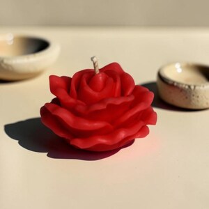 May include: A vibrant red candle shaped like a blooming rose with layered, curved petals and a single central wick. The wax flower is sculpted with realistic detail, capturing the organic form of a rose in full bloom, positioned upright.