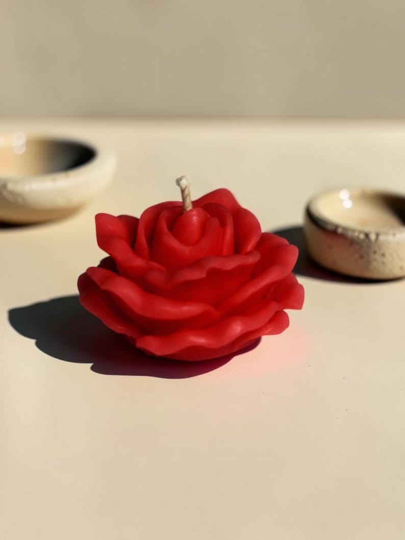 May include: A vibrant red candle shaped like a blooming rose with layered, curved petals and a single central wick. The wax flower is sculpted with realistic detail, capturing the organic form of a rose in full bloom, positioned upright.