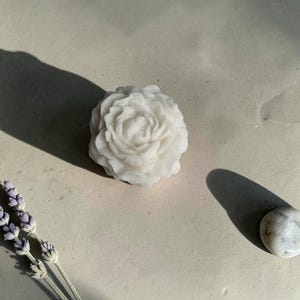 Pu&ograve; includere: Un sapone bianco modellato a forma di fiore di peonia in fiore con petali complessi e stratificati. Il sapone &egrave; posizionato accanto a un rametto di lavanda essiccata e a una piccola pietra bianca liscia, che proiettano ombre nette.