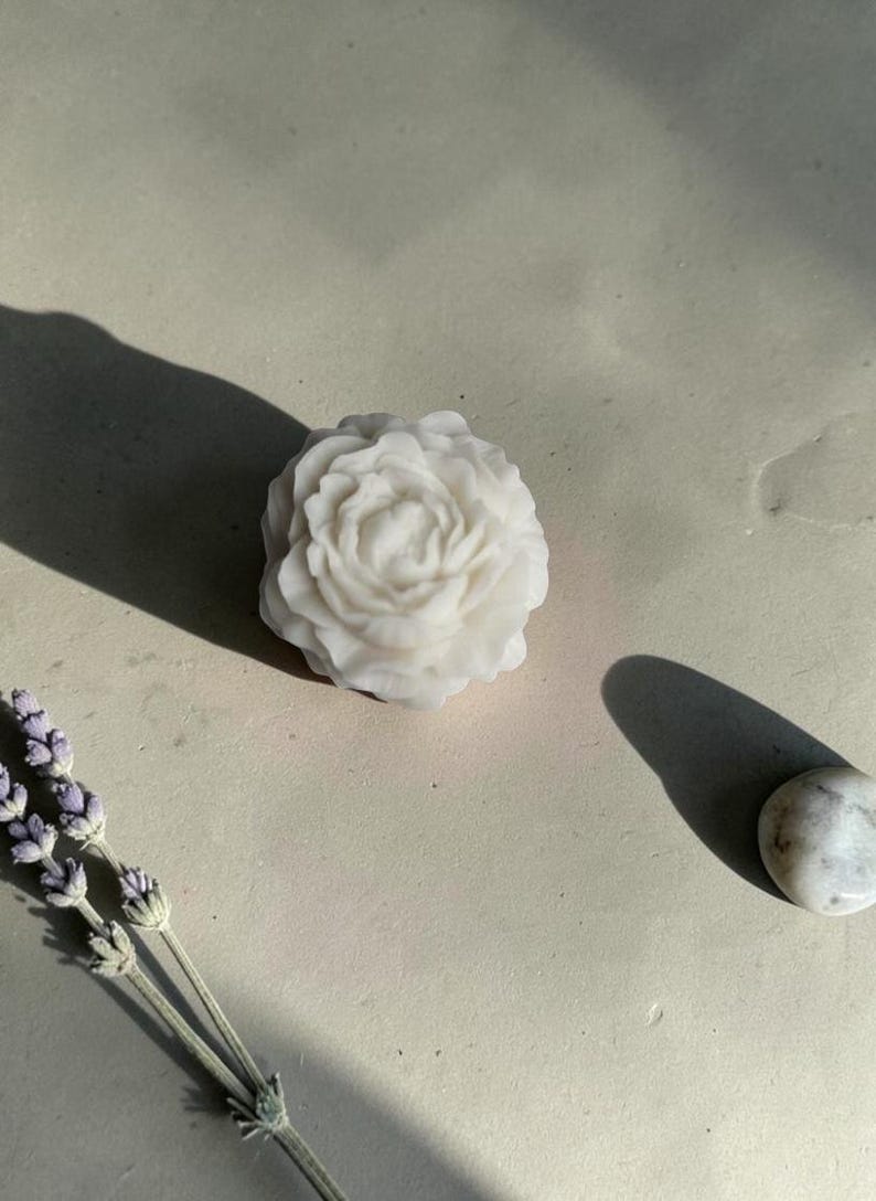 Pu&ograve; includere: Un sapone bianco modellato a forma di fiore di peonia in fiore con petali complessi e stratificati. Il sapone &egrave; posizionato accanto a un rametto di lavanda essiccata e a una piccola pietra bianca liscia, che proiettano ombre nette.