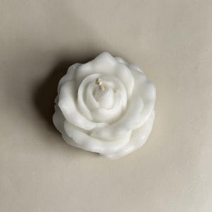 May include: A white wax candle shaped like a blooming rose with multiple layered petals. The candle features a single central cotton wick protruding from the center of the flower. The wax has a smooth, matte finish and a realistic floral structure.