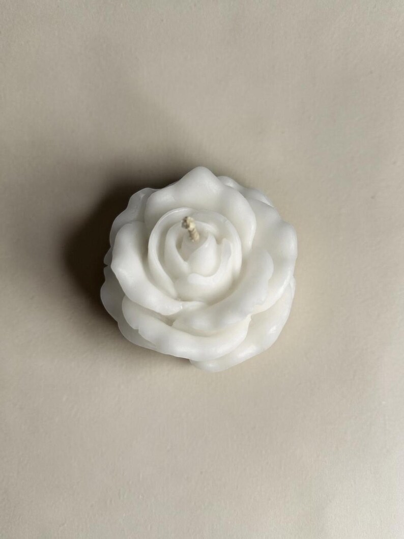 May include: A white wax candle shaped like a blooming rose with multiple layered petals. The candle features a single central cotton wick protruding from the center of the flower. The wax has a smooth, matte finish and a realistic floral structure.