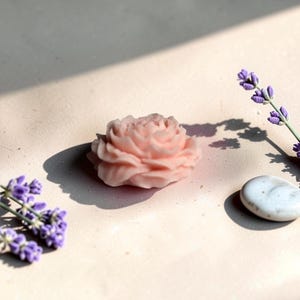 Pu&ograve; includere: Un sapone o una cera a forma di rosa color rosa pallido con petali stratificati e intricati. L'oggetto &egrave; posizionato accanto a due rametti di fiori di lavanda viola e a una singola pietra ovale bianca e liscia, che proiettano ombre nette.