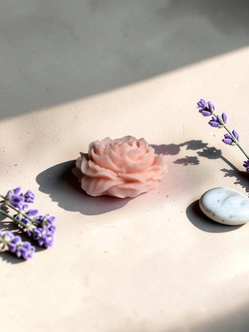 Pu&ograve; includere: Un sapone o una cera a forma di rosa color rosa pallido con petali stratificati e intricati. L'oggetto &egrave; posizionato accanto a due rametti di fiori di lavanda viola e a una singola pietra ovale bianca e liscia, che proiettano ombre nette.
