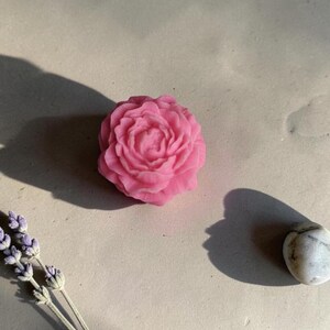 Pu&ograve; includere: Una saponetta rosa a forma di rosa con dettagli intricati di petali sovrapposti. Il sapone &egrave; posizionato accanto a un piccolo rametto di lavanda essiccata e a una singola pietra liscia maculata di bianco e grigio.