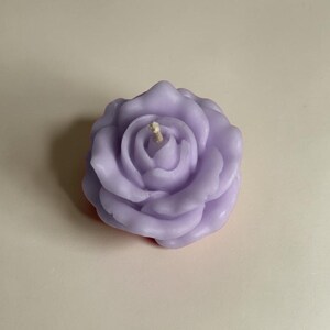 May include: A light purple candle shaped like a blooming rose with multiple layered petals and a single central cotton wick. The wax has a smooth, matte finish and is molded into a detailed floral structure with curved, overlapping petal edges.