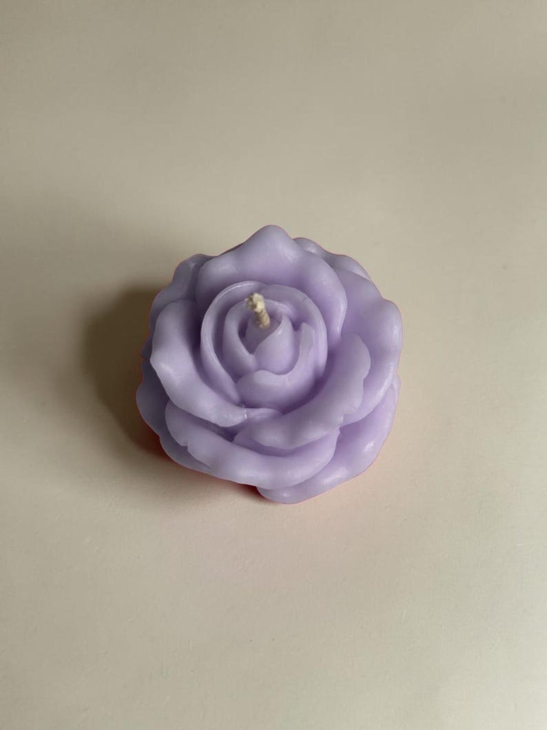 May include: A light purple candle shaped like a blooming rose with multiple layered petals and a single central cotton wick. The wax has a smooth, matte finish and is molded into a detailed floral structure with curved, overlapping petal edges.