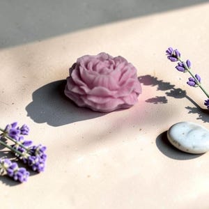 Pu&ograve; includere: Un sapone viola chiaro a forma di fiore di rosa in piena fioritura con dettagli intricati sui petali. Il sapone &egrave; posizionato accanto a due rametti di fiori di lavanda viola e una singola pietra bianca liscia di forma ovale su una superficie piana.