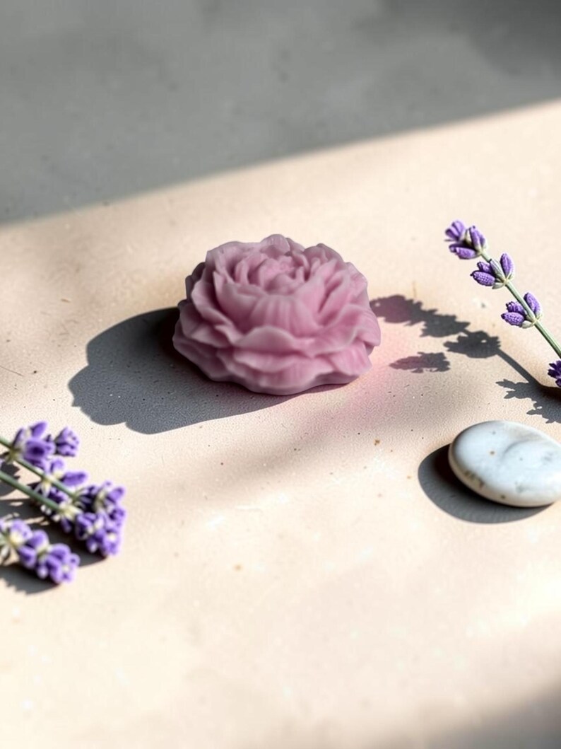Pu&ograve; includere: Un sapone viola chiaro a forma di fiore di rosa in piena fioritura con dettagli intricati sui petali. Il sapone &egrave; posizionato accanto a due rametti di fiori di lavanda viola e una singola pietra bianca liscia di forma ovale su una superficie piana.