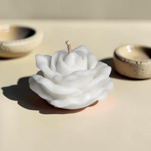 May include: A white wax candle shaped like a blooming rose with layered petals and a single centered wick. The candle has a smooth, matte finish and is positioned to show its full, rounded floral form with soft shadows cast on the surface.