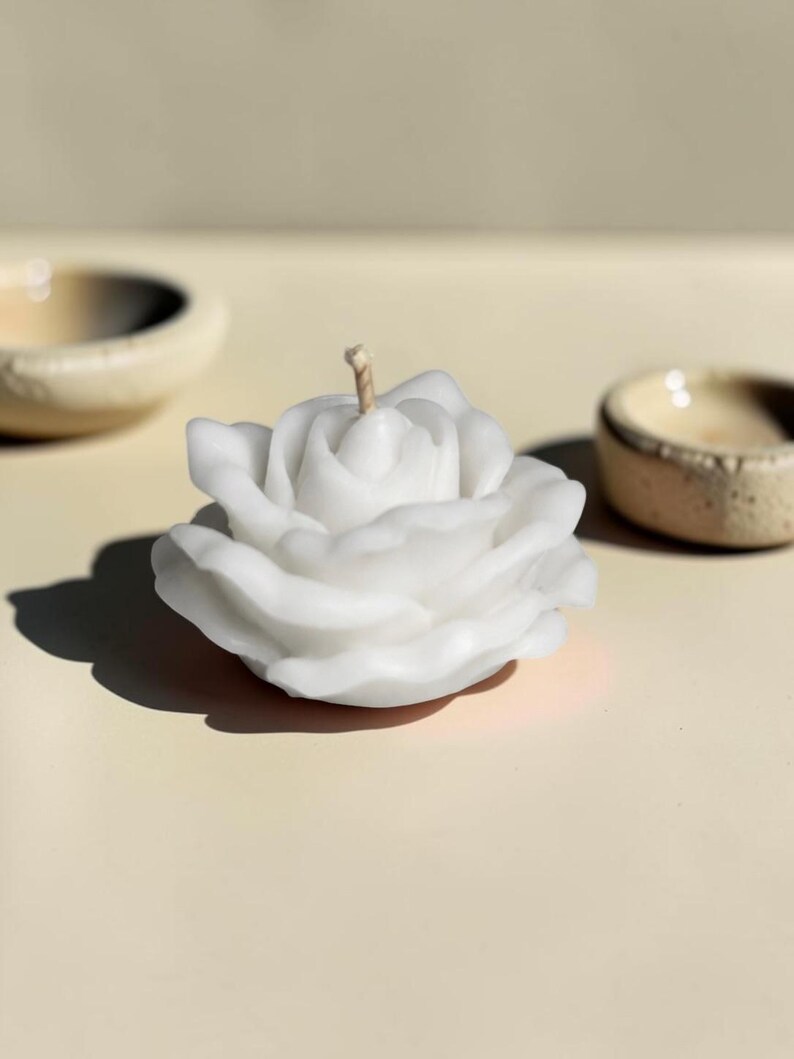 May include: A white wax candle shaped like a blooming rose with layered petals and a single centered wick. The candle has a smooth, matte finish and is positioned to show its full, rounded floral form with soft shadows cast on the surface.