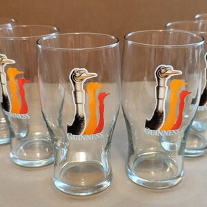 May include: Set of six clear pint glasses. Each glass features a logo of a bird with black, orange, and red accents, and the word "Guinness" below. The glasses are arranged in a semi-circle.