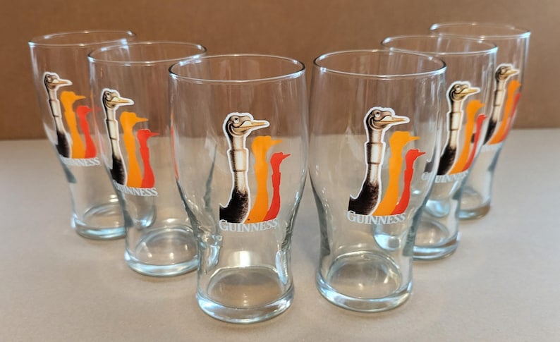 May include: Set of six clear pint glasses. Each glass features a logo of a bird with black, orange, and red accents, and the word "Guinness" below. The glasses are arranged in a semi-circle.