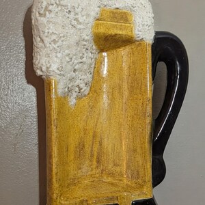 May include: A ceramic wall plaque shaped like a beer mug with a yellow body, a black handle and base, and a textured white foam top. The design features a glossy finish on the yellow and black sections, contrasting with the rough, porous white foam.